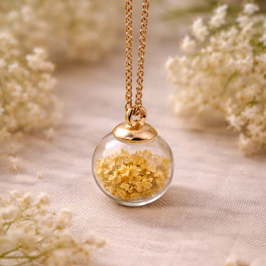 Gelbe Holunderblüten in einer Glasperle, an einer goldenen Kette und umringt von getrockneten Blumen